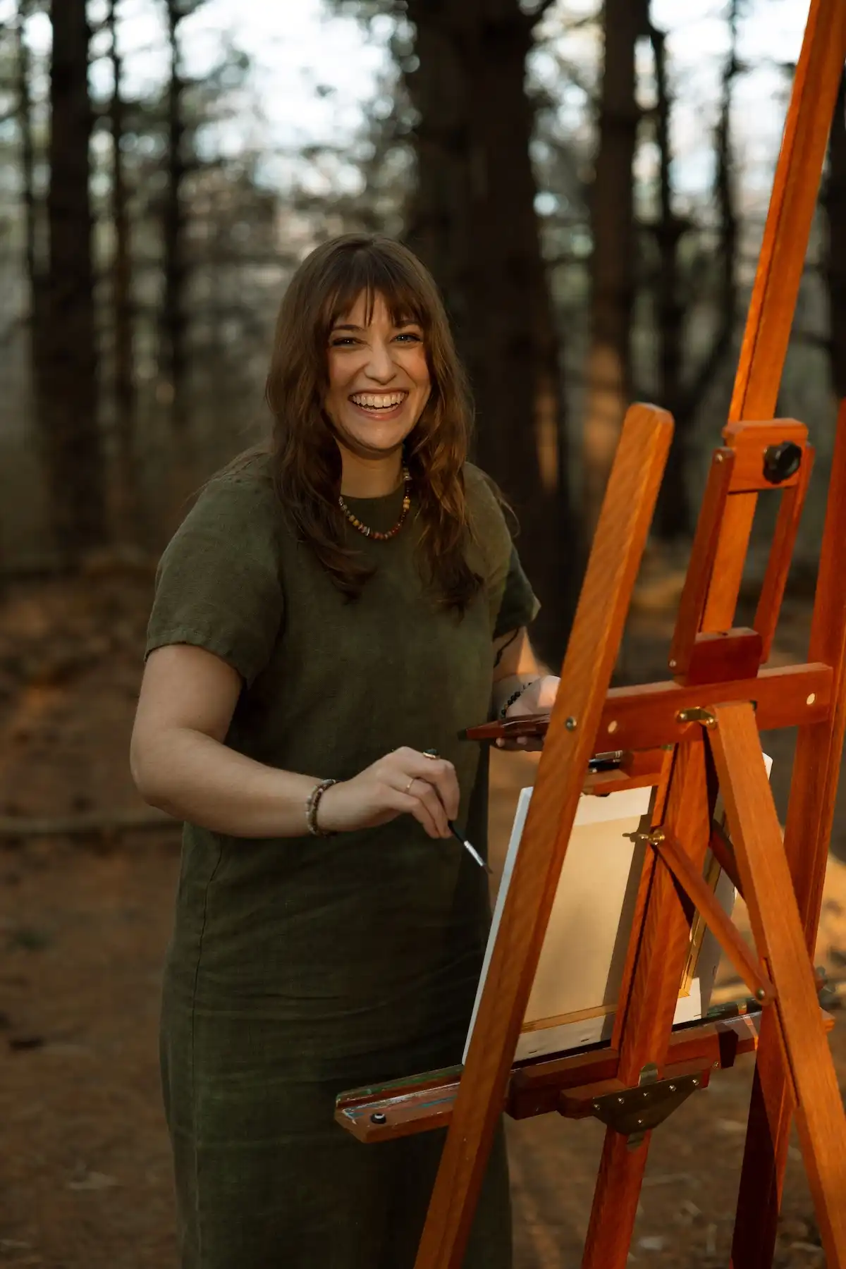Madeline Holloway smiling while painting at an easel in a pine forest
